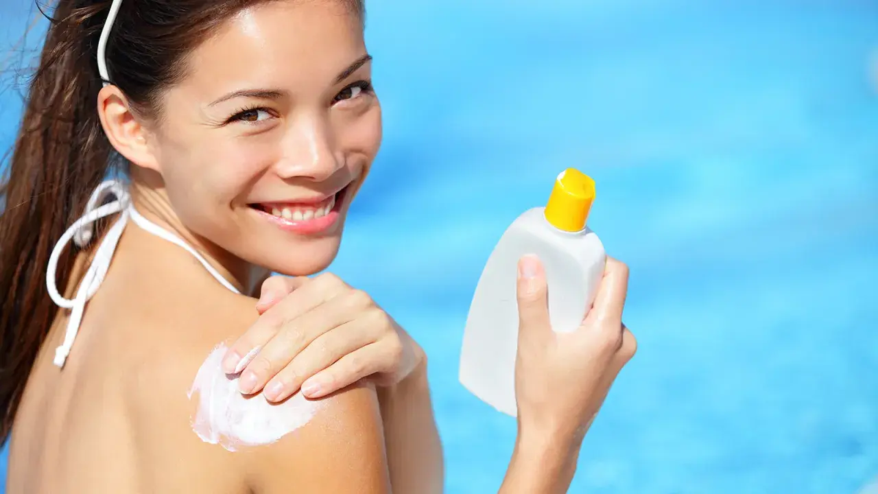 Donna sorridente applica crema solare sulla spalla, con una bottiglia in mano. Scopri che protezione solare usare per la tua pelle.