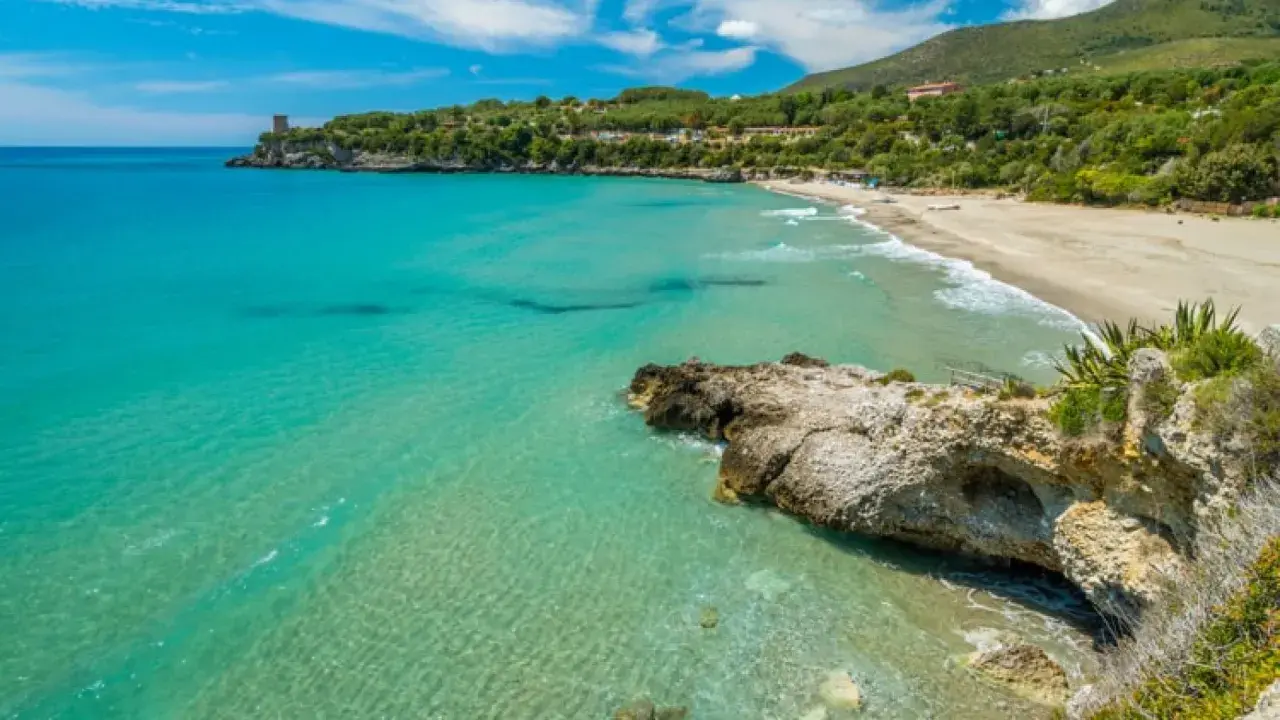 Spiaggia dorata e mare cristallino nel Cilento, con una torre antica su una scogliera e colline verdi sullo sfondo.