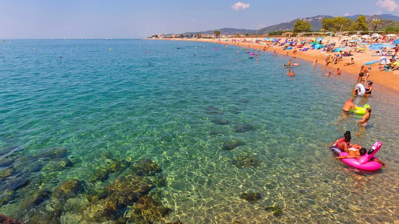 Spiaggia affollata a Santa Susanna, Spagna, con persone che nuotano e si rilassano in un mare cristallino.