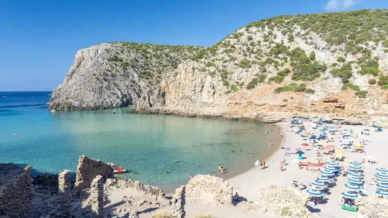 Spiaggia affollata con ombrelloni colorati, acque cristalline e scogliere verdi. Perfetta per vacanze al mare economiche e rilassanti.