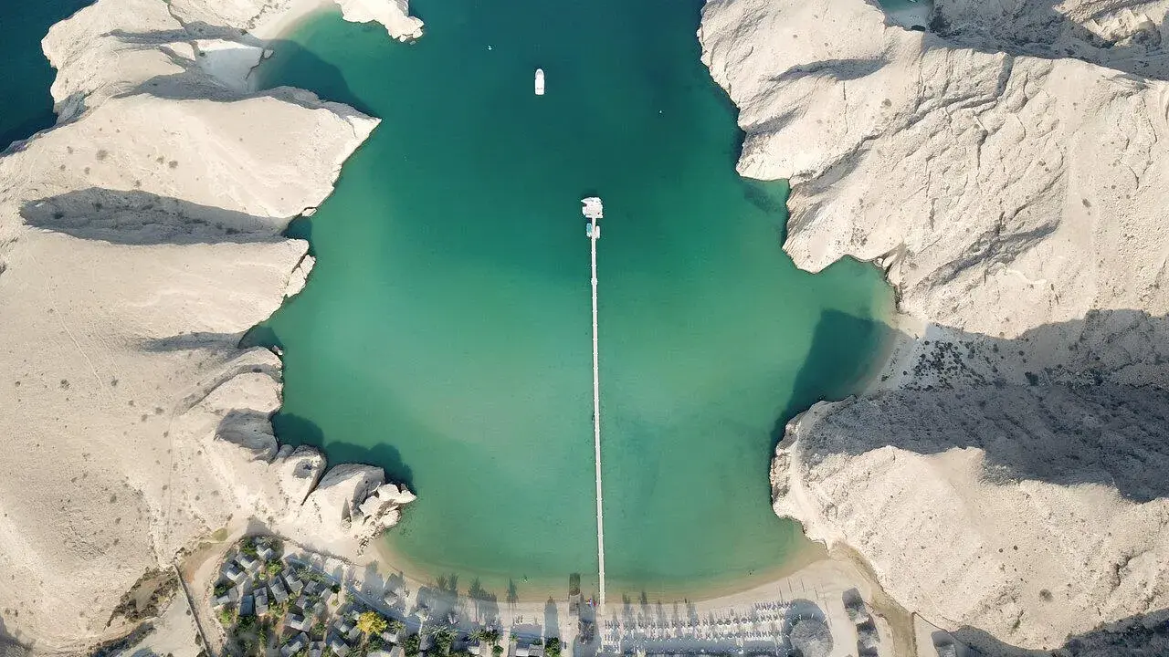 Vista aerea di un resort in Oman con bambini che giocano sulla spiaggia. Acque turchesi, montagne rocciose e un lungo pontile.