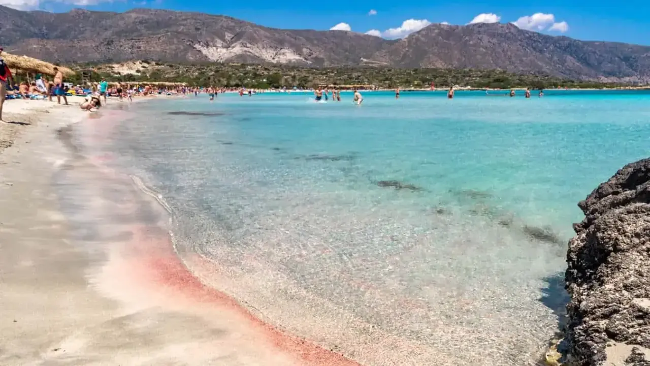 Spiaggia rosa di Creta con bambini che giocano nell'acqua cristallina e montagne sullo sfondo.