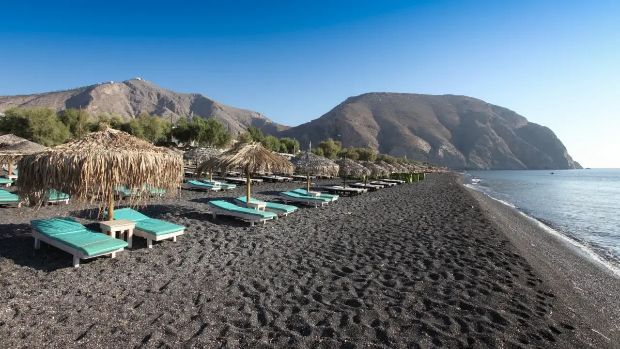 Santorini spiagge di sabbia nera con lettini e ombrelloni di paglia, montagne sullo sfondo e mare calmo.