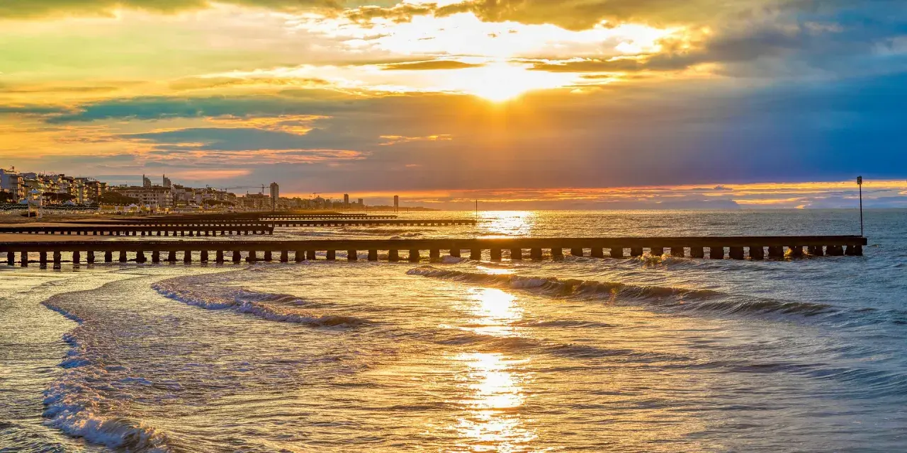 Tramonto dorato sul mare veneto, con moli che si estendono nell'acqua e una città all'orizzonte.