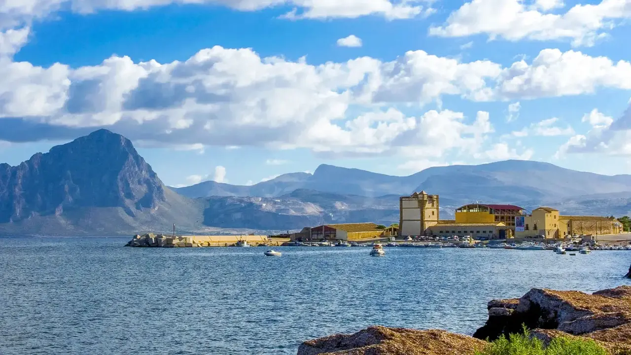 La tonnara di Bonagia, con il suo porto e le montagne sullo sfondo, sotto un cielo azzurro e nuvoloso.