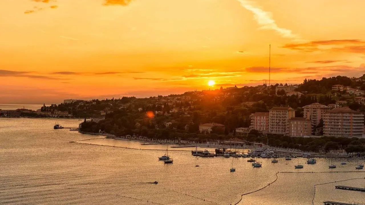 Tramonto dorato sul mare in Slovenia, con un porto pieno di barche e una costa verdeggiante. Immagina i bambini che giocano felici sulla spiaggia.
