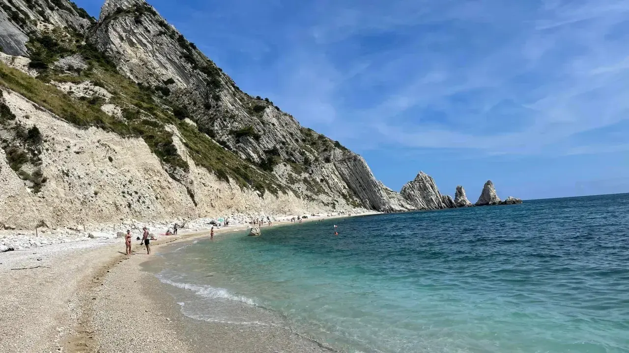 Spiaggia della Riviera del Conero con bambini che giocano e si bagnano, scogliere bianche e mare azzurro.