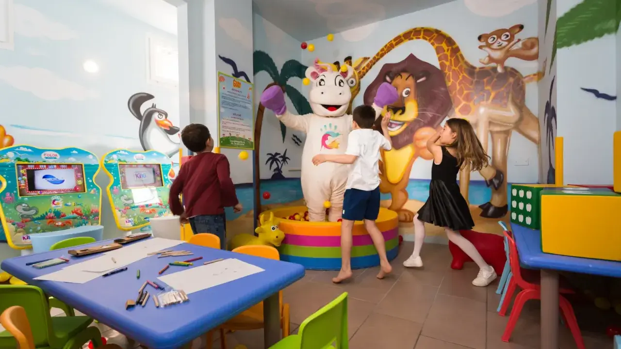 Bambini giocano con un personaggio in costume da ippopotamo in una stanza colorata, un'area giochi per famiglie al Mokambo Shore Easy Family Hotel.