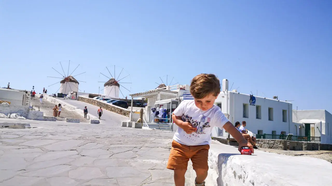 Un bambino gioca con una macchinina rossa a Mykonos, con i mulini a vento sullo sfondo. Vacanze a Mykonos con bambini.