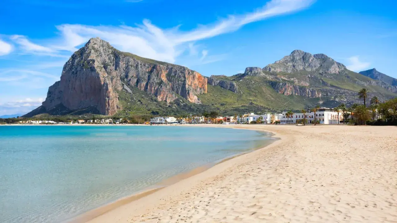 Spiaggia dorata e mare cristallino, un angolo di paradiso in Sicilia dove andare al mare. Montagne imponenti sullo sfondo.