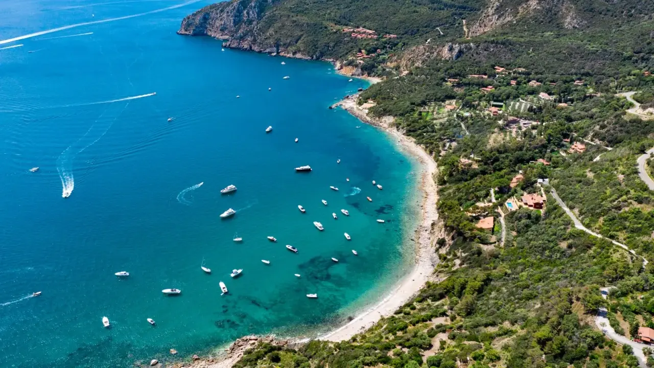 Baia incantevole con mare bello toscana, acque cristalline e barche che navigano. La costa è verdeggiante, con ville sparse tra gli alberi.