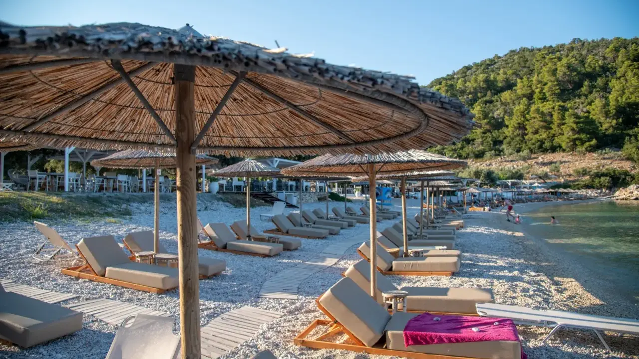Alonissos spiagge: lettini e ombrelloni di paglia su una spiaggia di ciottoli, con il mare cristallino e la collina verde sullo sfondo.