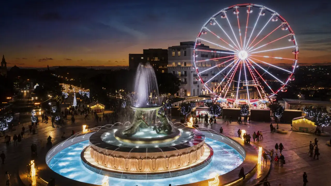 Una piazza illuminata a festa a Malta a Natale, con una fontana scintillante e una ruota panoramica che domina lo skyline al tramonto.