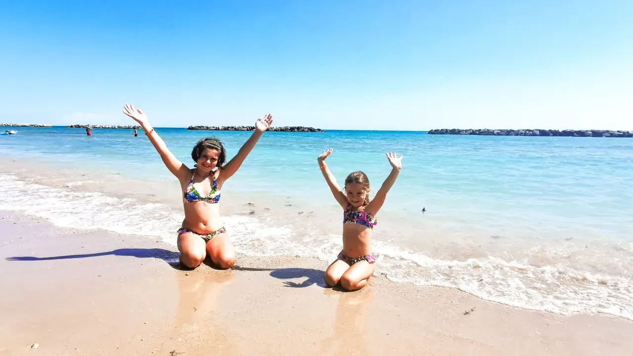 Due bambine felici sulla spiaggia del villaggio Riva Verde, Marche, con le braccia alzate verso il cielo azzurro.