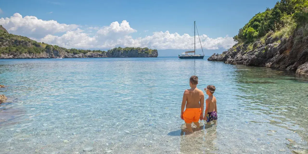 Due bambini esplorano le acque cristalline del Cilento, tra le sue spiagge perfette per famiglie. Una barca a vela all'orizzonte completa il quadro idilliaco.