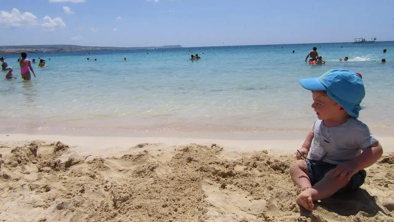 Bambino con cappellino blu gioca sulla sabbia a Cipro, mentre altri si divertono in mare.