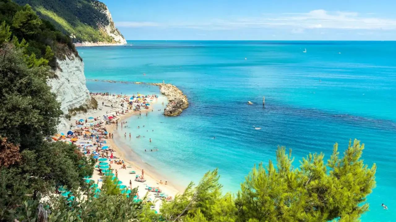 Spiaggia affollata con ombrelloni colorati e gente che nuota nel mare cristallino dell'Abruzzo. Scogliera bianca e vegetazione rigogliosa incorniciano questo angolo di paradiso.