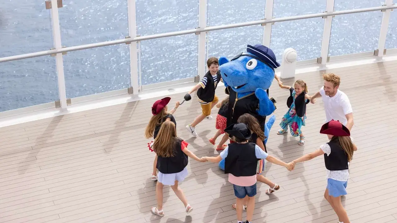Bambini giocano in cerchio con un personaggio blu su una nave, con la costa toscana sullo sfondo.