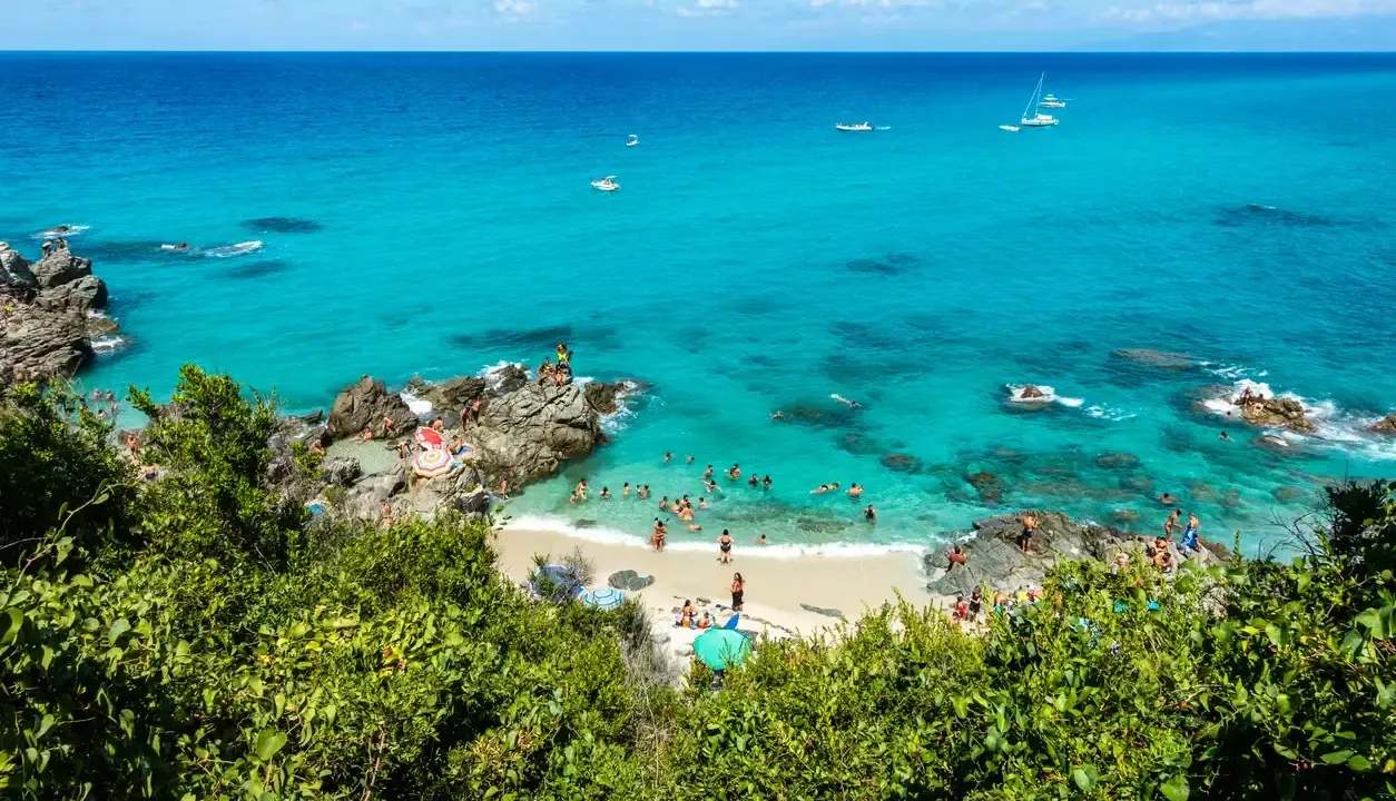 Spiaggia affollata con gente che nuota e prende il sole, barche in mare e vegetazione lussureggiante. Un angolo di paradiso del **calabria mare**.