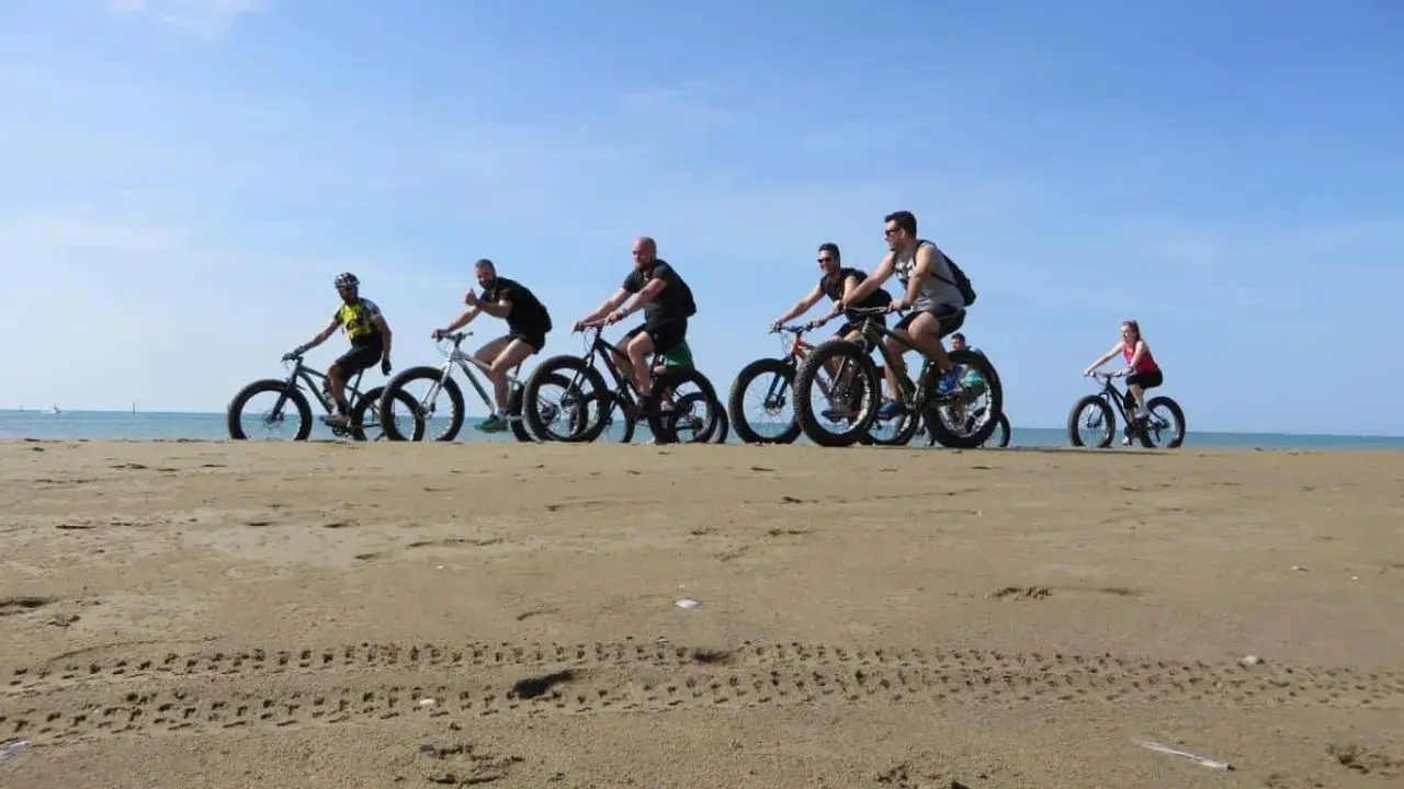 Gruppo di ciclisti su fat bike lungo la ciclabile di Caorle, sulla sabbia dorata con il mare sullo sfondo.
