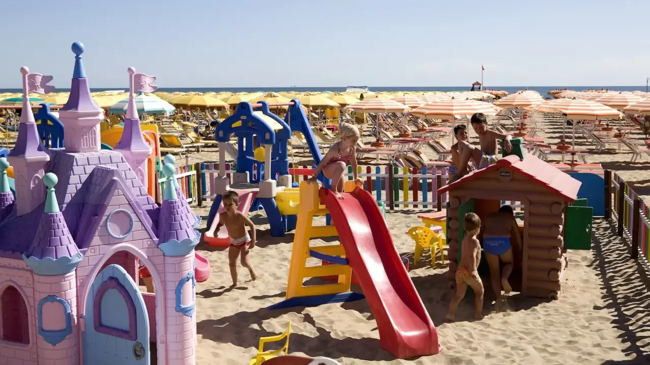 Bambini giocano in un'area giochi sulla spiaggia di Rimini, tra castelli rosa, scivoli e casette di legno. Perfetto per scoprire cosa fare a Rimini con i bambini!