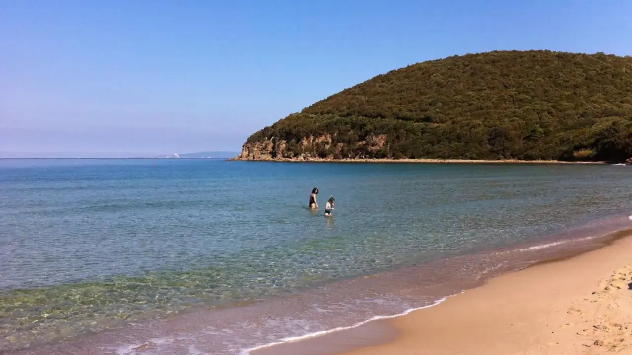 Spiaggia della Maremma con due bambini che giocano nell'acqua cristallina, sotto un cielo azzurro e una collina verdeggiante.