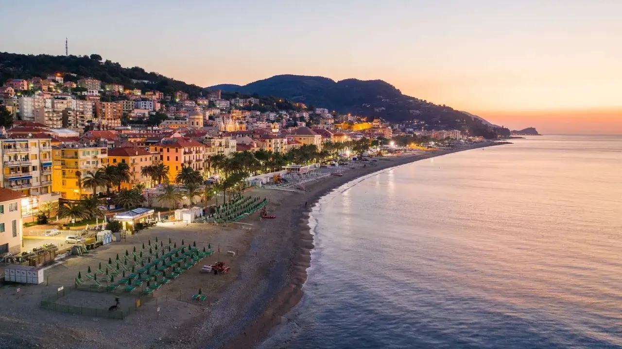 Alba sulla spiaggia finale ligure, con file di ombrelloni verdi e palme che si affacciano sul mare calmo.