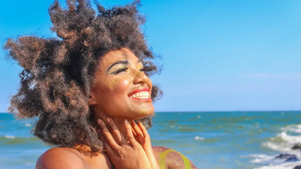 Donna con capelli afro e trucco scintillante si gode il sole in spiaggia. Sembra che ci si abbronza con il trucco, creando un look festoso.