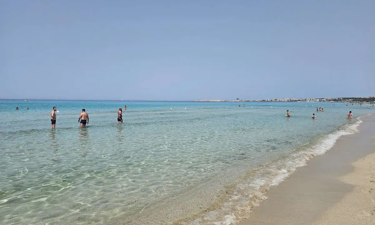 Persone in piedi nell'acqua cristallina, con la costa di Gallipoli sullo sfondo. Un perfetto giorno di mare.