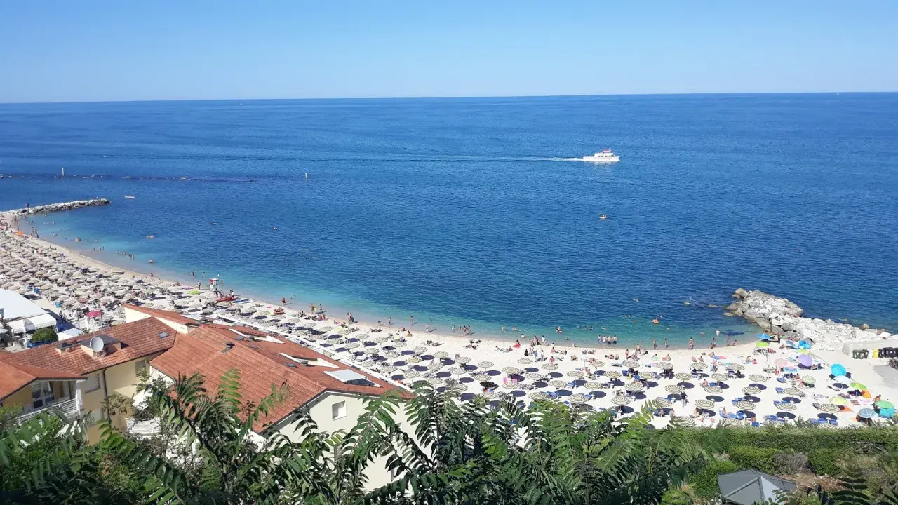 Le spiagge di Numana, con ombrelloni colorati e gente che si gode il sole, bagnate da un mare azzurro e calmo. Una barca solca l'orizzonte.