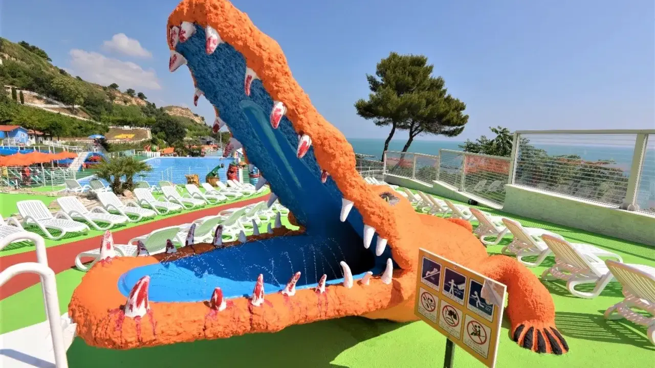 Un gigantesco coccodrillo arancione con la bocca spalancata, un divertente scivolo acquatico, si affaccia su una piscina e lettini bianchi, con una rigogliosa riva verde fermo sullo sfondo.