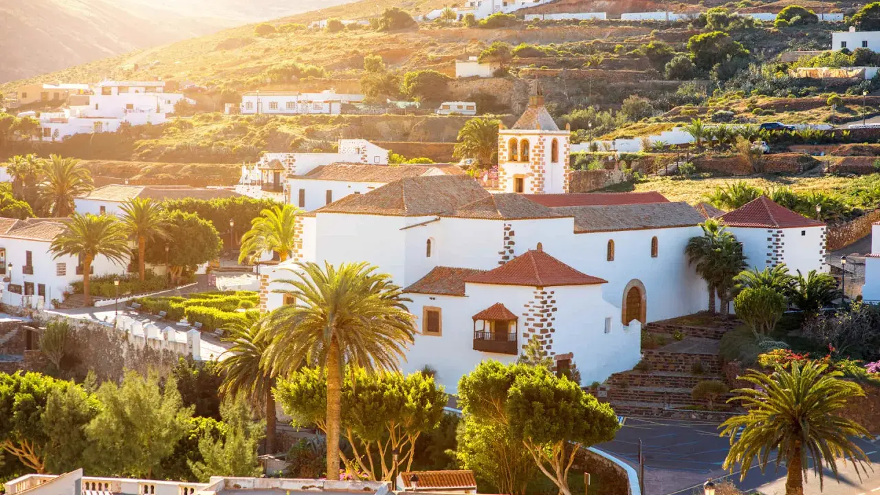 Villaggi turistici a Fuerteventura, con case bianche, palme e una chiesa con campanile, immersi nel sole dorato del tramonto.