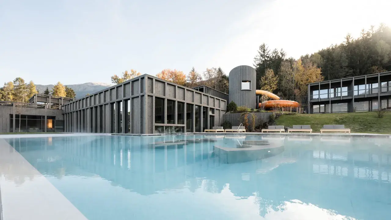 Piscina esterna con acqua fumante e scivoli acquatici arancioni al Aki Family Resort Plose. Architettura moderna in legno e montagne sullo sfondo.