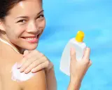 Donna sorridente applica crema solare sulla spalla, con una bottiglia in mano. Scopri che protezione solare usare per la tua pelle.
