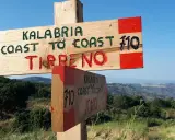 Cartello in legno indica "Kalabria Coast to Coast" verso il Tirreno e lo Jonio, con sfondo di colline e mare.