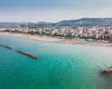 Vista aerea di San Benedetto del Tronto: una lunga spiaggia con ombrelloni colorati e il mare turchese, con dighe frangiflutti che proteggono la costa.