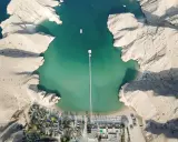 Vista aerea di un resort in Oman con bambini che giocano sulla spiaggia. Acque turchesi, montagne rocciose e un lungo pontile.