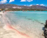 Spiaggia rosa di Creta con bambini che giocano nell'acqua cristallina e montagne sullo sfondo.