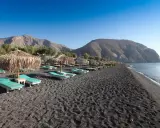 Santorini spiagge di sabbia nera con lettini e ombrelloni di paglia, montagne sullo sfondo e mare calmo.