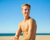 Giovane uomo sorridente sulla spiaggia, con la pelle dorata come se con l'autoabbronzante potesse prendere il sole senza rischi.