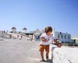 Un bambino gioca con una macchinina rossa a Mykonos, con i mulini a vento sullo sfondo. Vacanze a Mykonos con bambini.