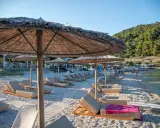 Alonissos spiagge: lettini e ombrelloni di paglia su una spiaggia di ciottoli, con il mare cristallino e la collina verde sullo sfondo.
