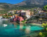 Case colorate affacciate su acque cristalline, un sogno di spiagge Grecia. Barche a vela e colline verdi completano il quadro.