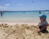 Bambino con cappellino blu gioca sulla sabbia a Cipro, mentre altri si divertono in mare.