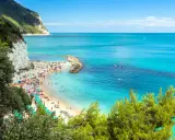 Spiaggia affollata con ombrelloni colorati e gente che nuota nel mare cristallino dell'Abruzzo. Scogliera bianca e vegetazione rigogliosa incorniciano questo angolo di paradiso.