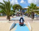 Accesso facilitato per tutti nei villaggi in Abruzzo sul mare. Un uomo in carrozzina elettrica percorre una passerella blu sulla sabbia, tra palme rigogliose e un edificio con la scritta "Welcome".
