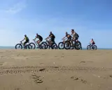 Gruppo di ciclisti su fat bike lungo la ciclabile di Caorle, sulla sabbia dorata con il mare sullo sfondo.