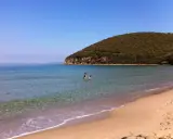 Spiaggia della Maremma con due bambini che giocano nell'acqua cristallina, sotto un cielo azzurro e una collina verdeggiante.