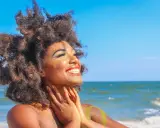Donna con capelli afro e trucco scintillante si gode il sole in spiaggia. Sembra che ci si abbronza con il trucco, creando un look festoso.