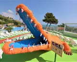 Un gigantesco coccodrillo arancione con la bocca spalancata, un divertente scivolo acquatico, si affaccia su una piscina e lettini bianchi, con una rigogliosa riva verde fermo sullo sfondo.